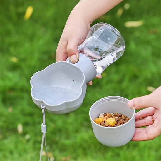 Démonstration de la gourde Pawfect 2 en 1 grise : une main verse de l'eau dans le réceptacle supérieur tandis que l'autre tient le compartiment inférieur rempli de croquettes et de friandises sur un fond d'herbe verte.