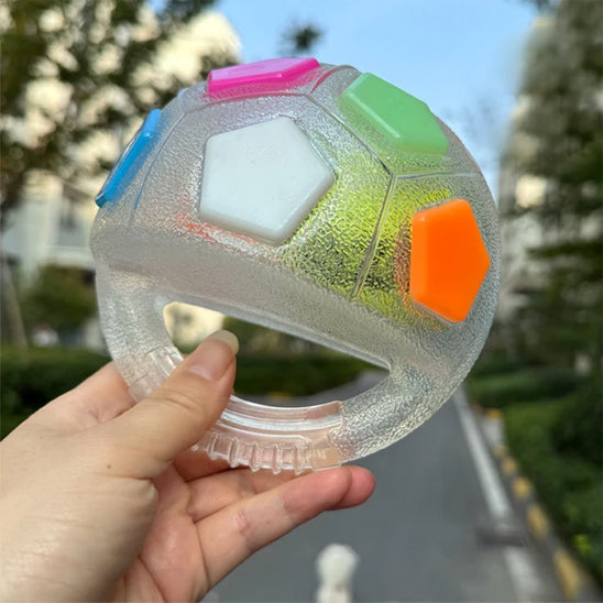 Balle de jeu transparente pour chien en forme de ballon de football américain, dotée d'une poignée centrale intégrée et de facettes colorées (rose, vert, orange, blanc). Une main tient le jouet en extérieur.