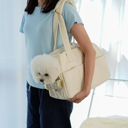 Gros plan de profil sur une femme portant à l'épaule le sac de transport en coton beige, d'où dépasse la tête d'un petit chien Bichon blanc.