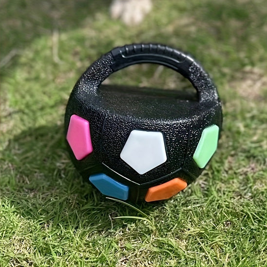 Jouet pour chien en forme de ballon de football américain de couleur noire, posé sur une pelouse verte. Le jouet possède une poignée intégrée sur le dessus et des facettes géométriques colorées en rose, blanc, vert, orange et bleu.